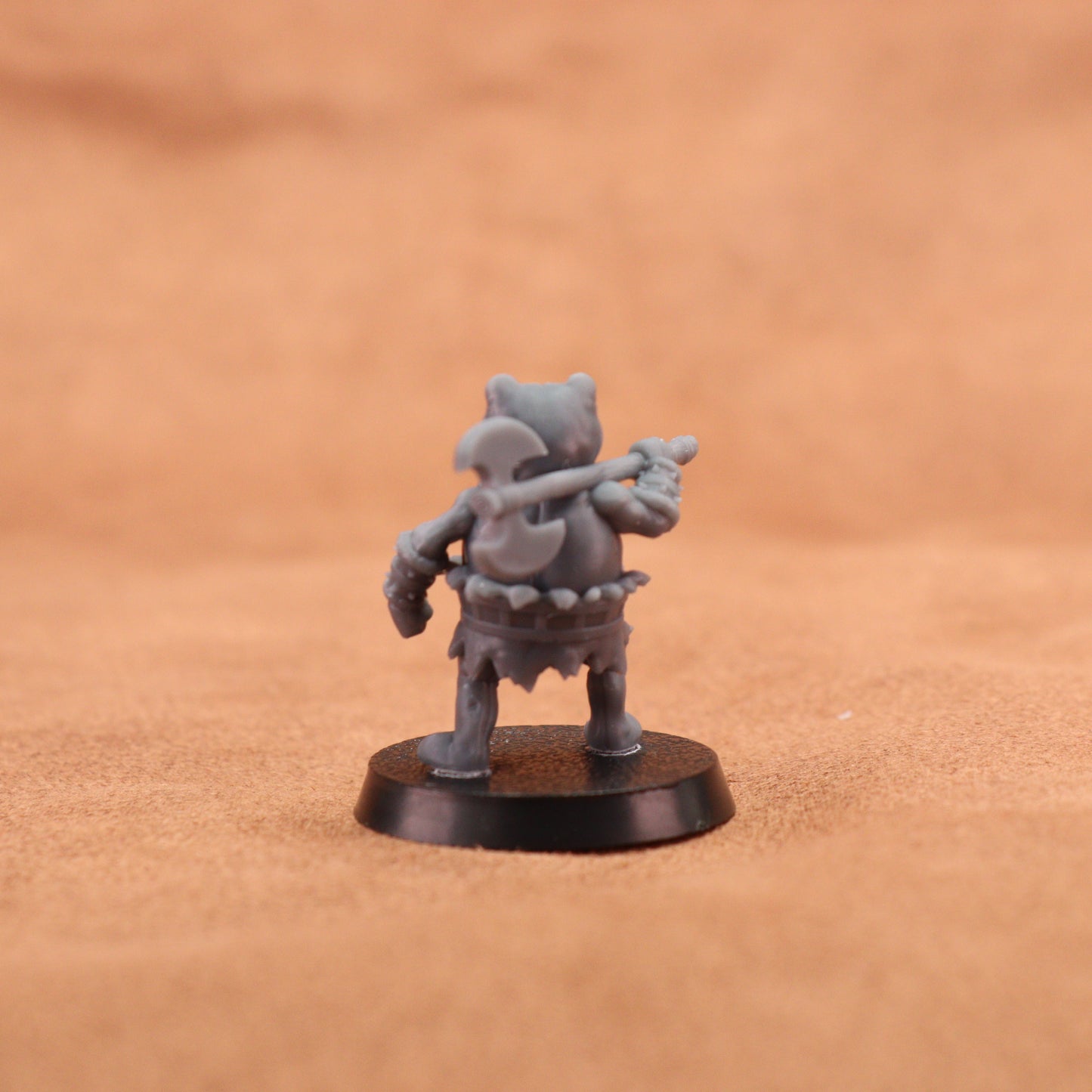 A small unpainted gray fantasy miniature figurine of a warrior holding a mace, standing on a round black base against a warm brown textured background.