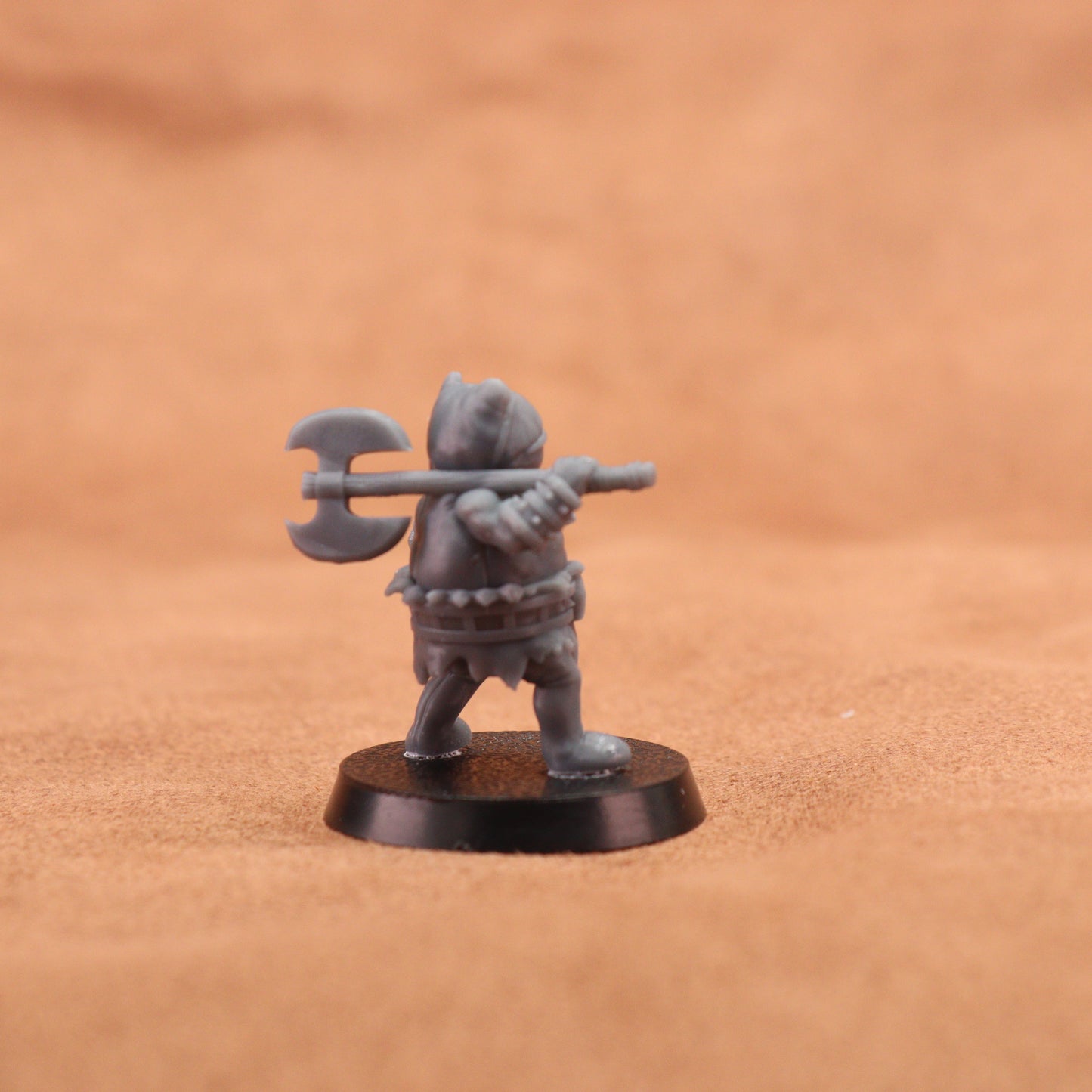 A small unpainted gray fantasy warrior miniature figurine holding a double-headed axe, standing on a round black base against a warm brown textured background.