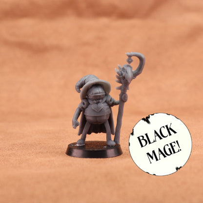 A small unpainted gray fantasy miniature figurine of a wizard holding a staff, standing on a round base against a warm brown textured background. A white circular sticker with the text 'BLACK MAGE!' is placed to the right.