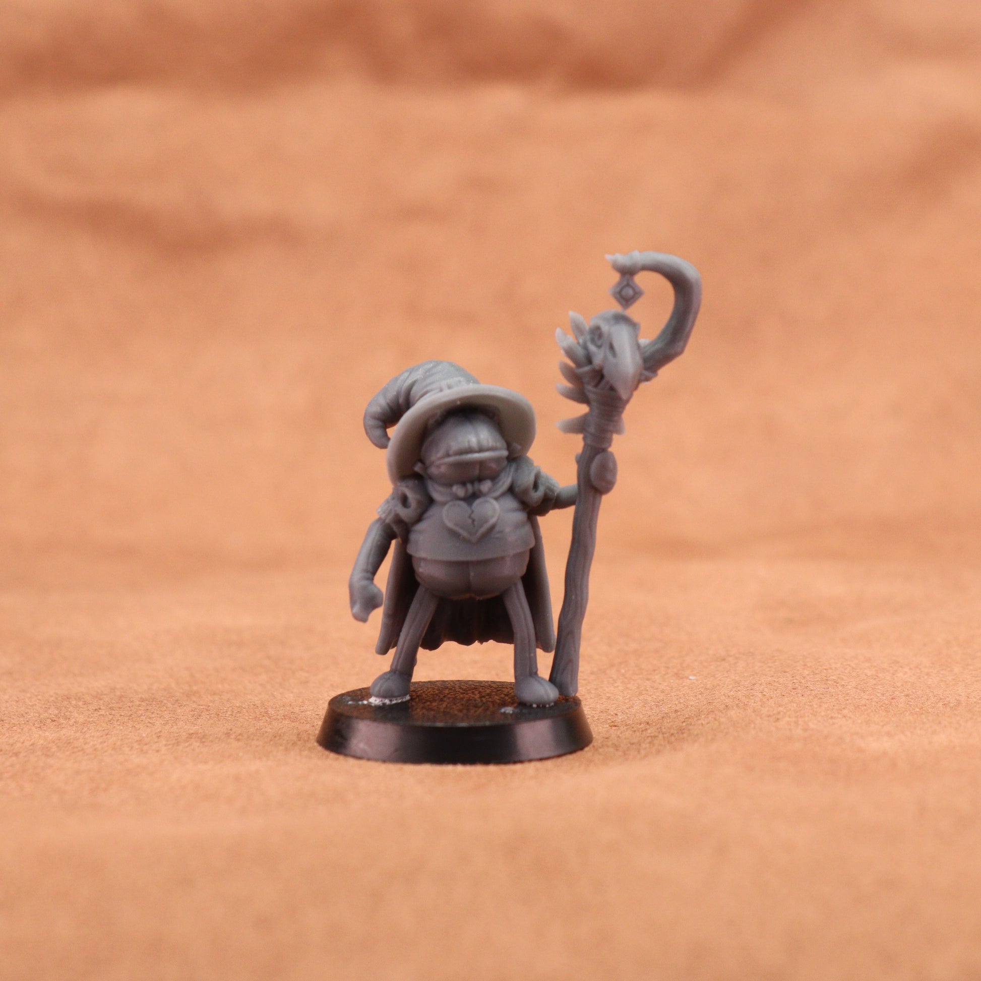 A small unpainted gray fantasy miniature figurine of a wizard holding a staff, standing on a round black base against a warm brown textured background.