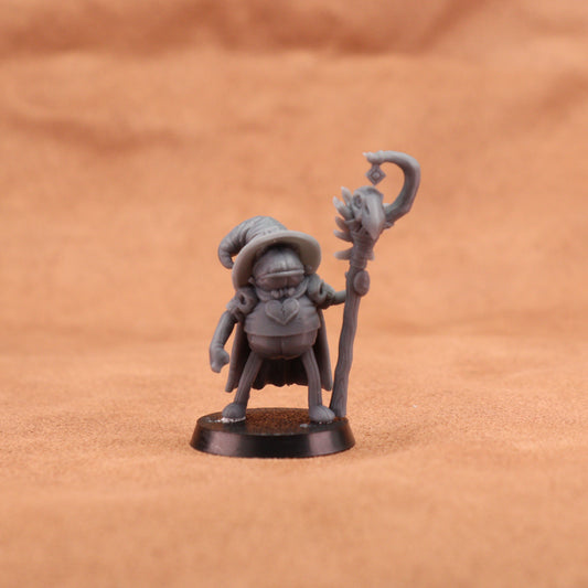 A small unpainted gray fantasy miniature figurine of a wizard holding a staff, standing on a round black base against a warm brown textured background.