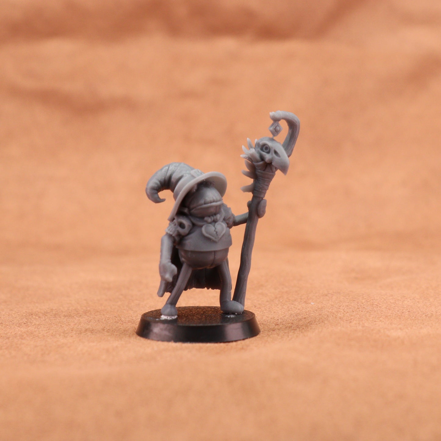 A small unpainted gray fantasy miniature figurine of a wizard-like character holding a staff topped with a skull, standing on a round black base against a warm brown textured background.