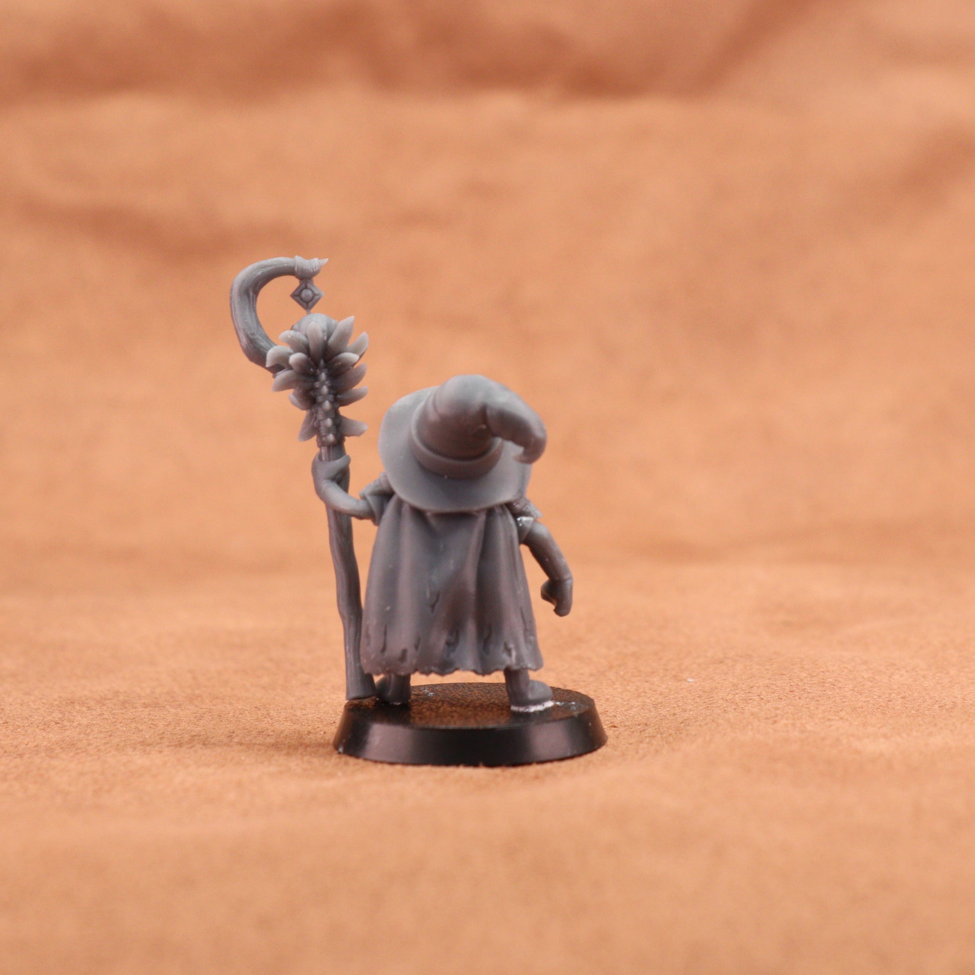 A small unpainted gray fantasy wizard miniature figurine holding a staff, standing on a round black base against a textured brown background.