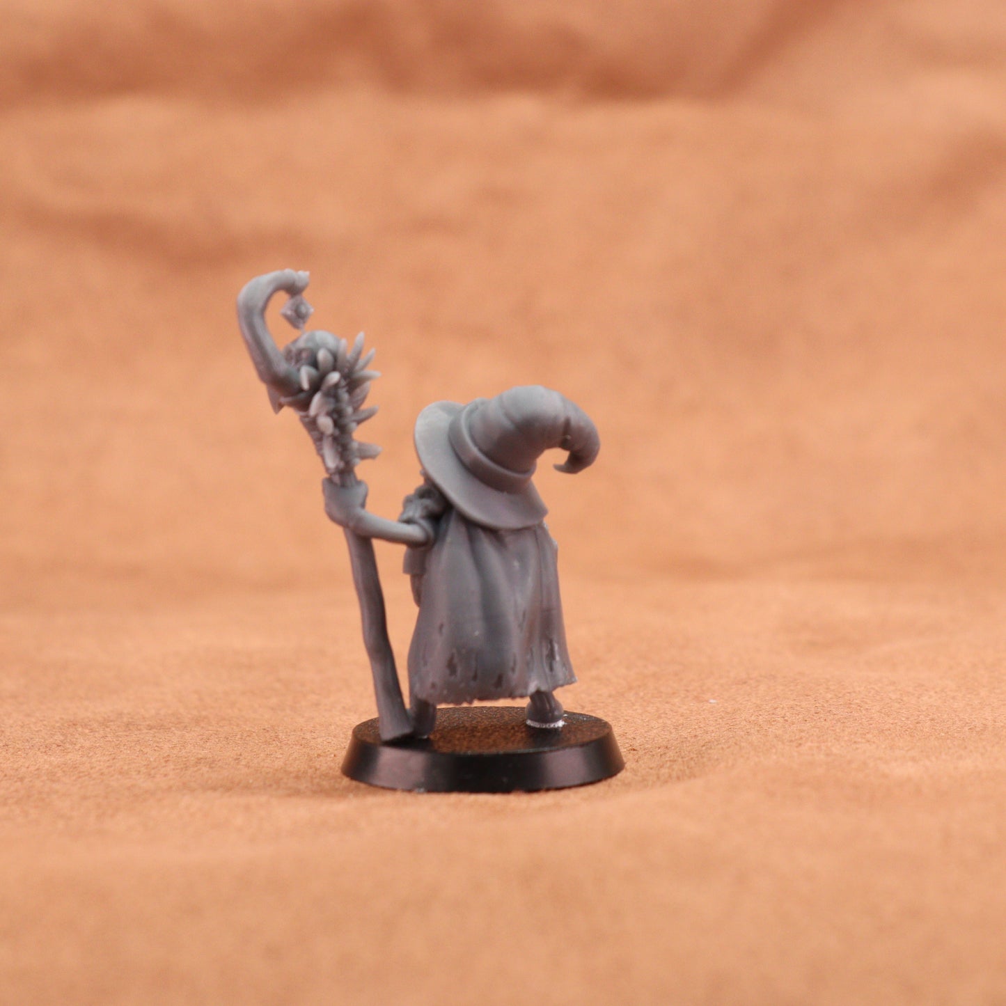 A small unpainted fantasy miniature figurine of a wizard or elf-like character holding a staff, standing on a round black base against a warm tan textured background.