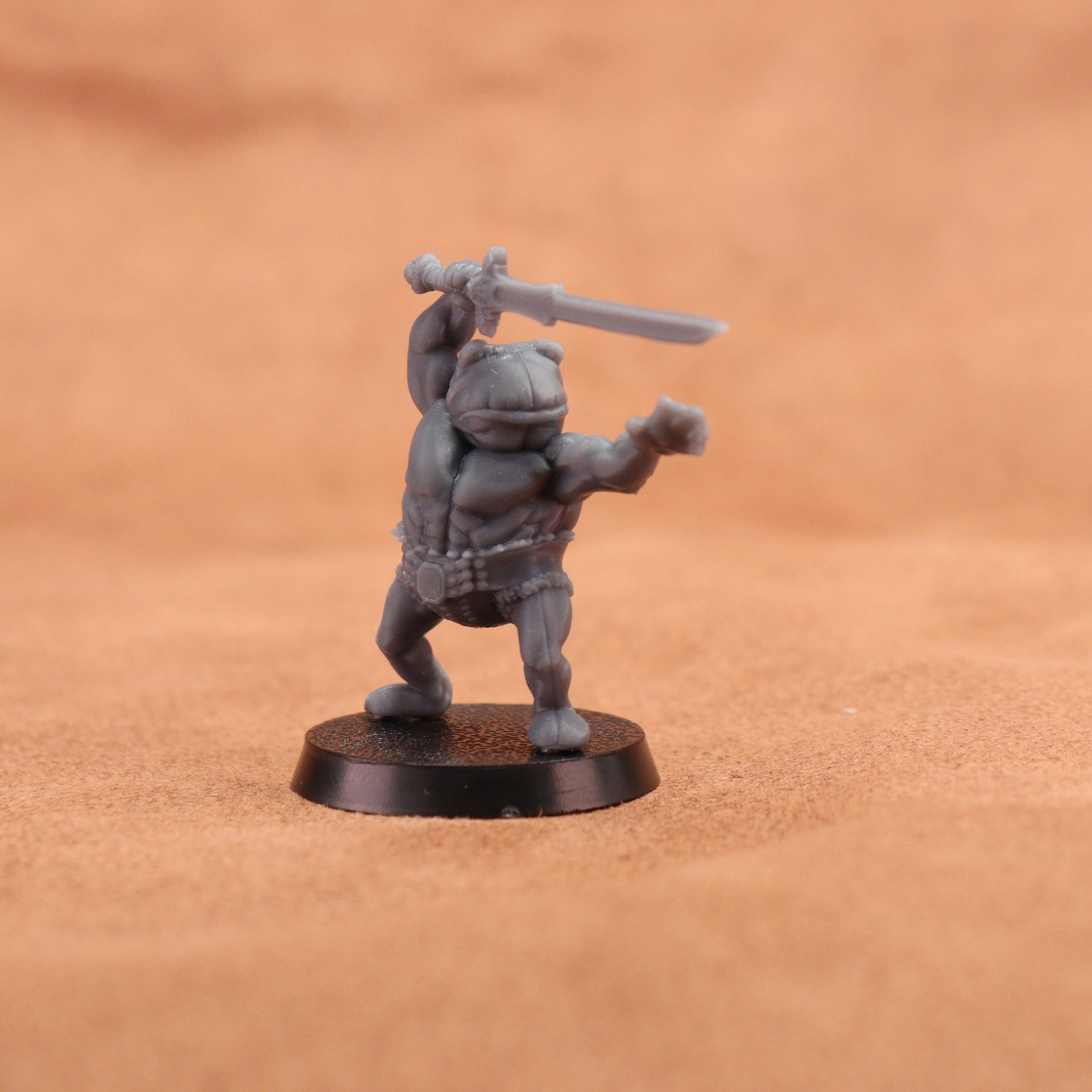 A small unpainted gray fantasy miniature figurine of a barbarian holding a sword, standing on a round black base against a warm brown textured background.