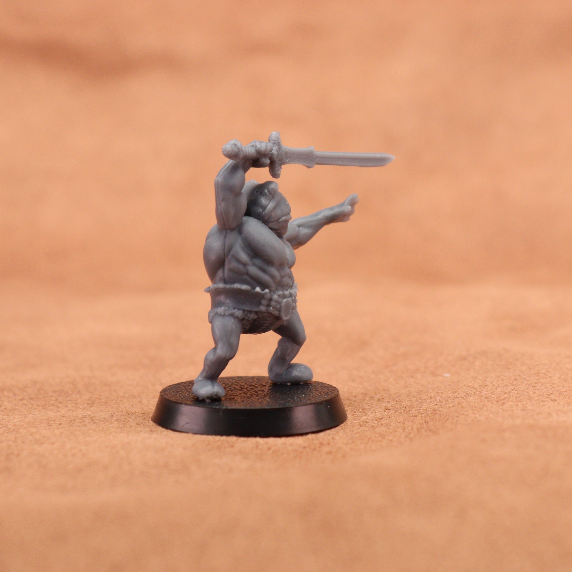 A small unpainted gray fantasy miniature figurine of a barbarian holding a sword, standing on a round black base against a warm brown textured background.
