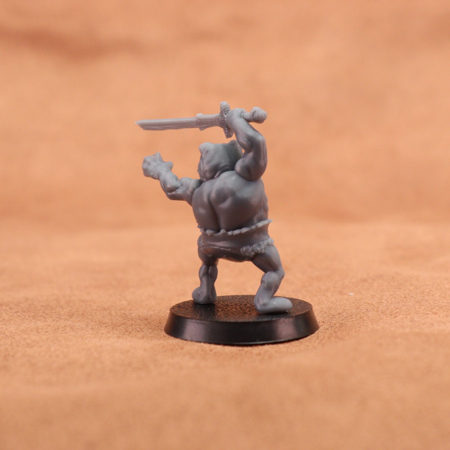A small unpainted gray fantasy miniature figurine of a barbarian holding a sword, standing on a round black base against a warm brown textured background.