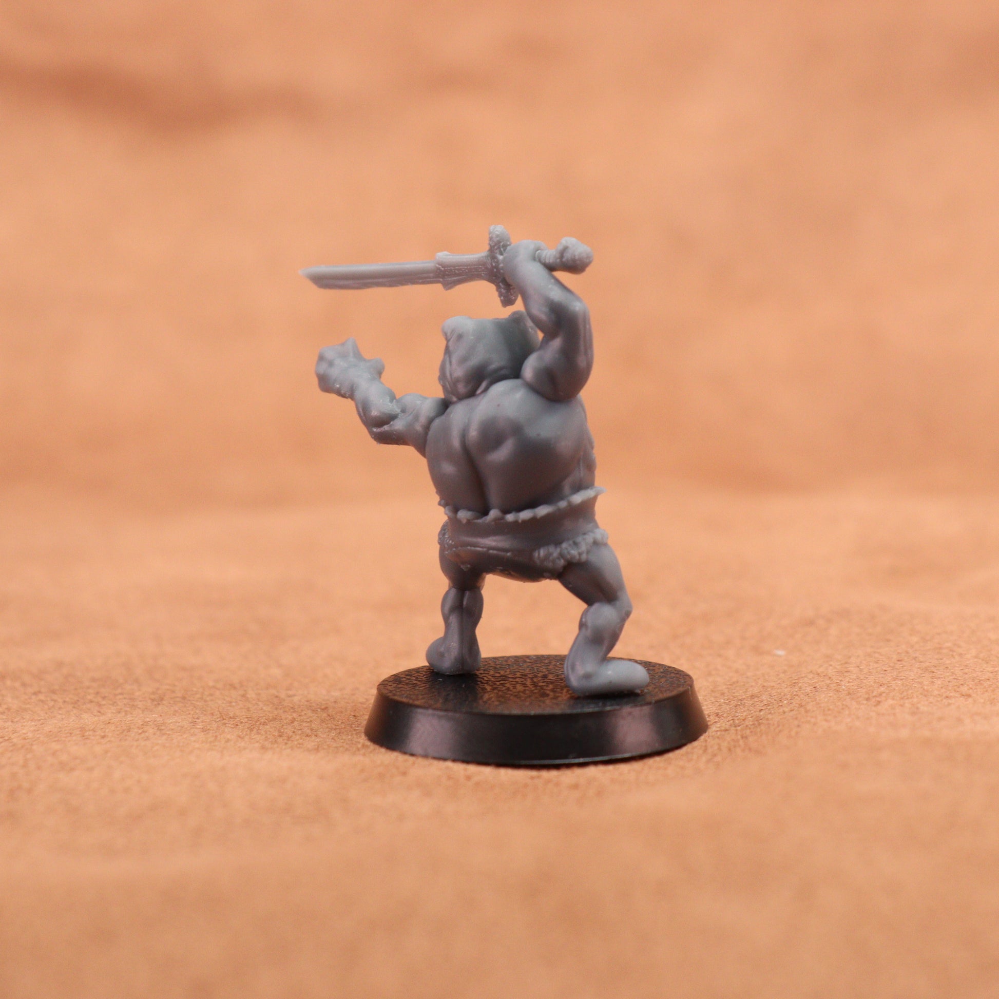 A small unpainted gray fantasy miniature figurine of a barbarian holding a sword, standing on a round black base against a warm brown textured background.