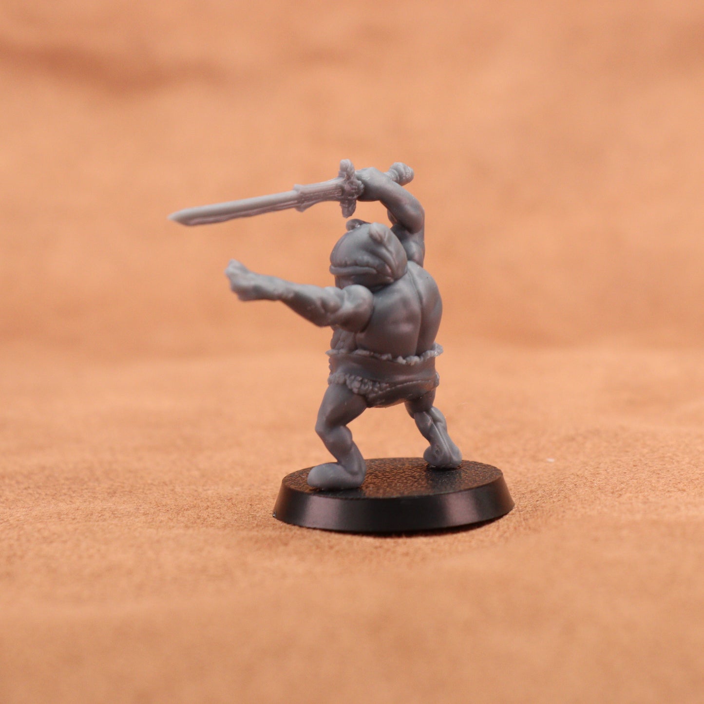 A small unpainted gray fantasy miniature figurine of a barbarian holding a sword, standing on a round black base against a warm brown textured background.