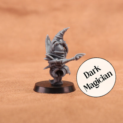 A small unpainted fantasy miniature figurine labeled 'Dark Magician' in a circular badge, standing on a round black base against a warm tan textured background.