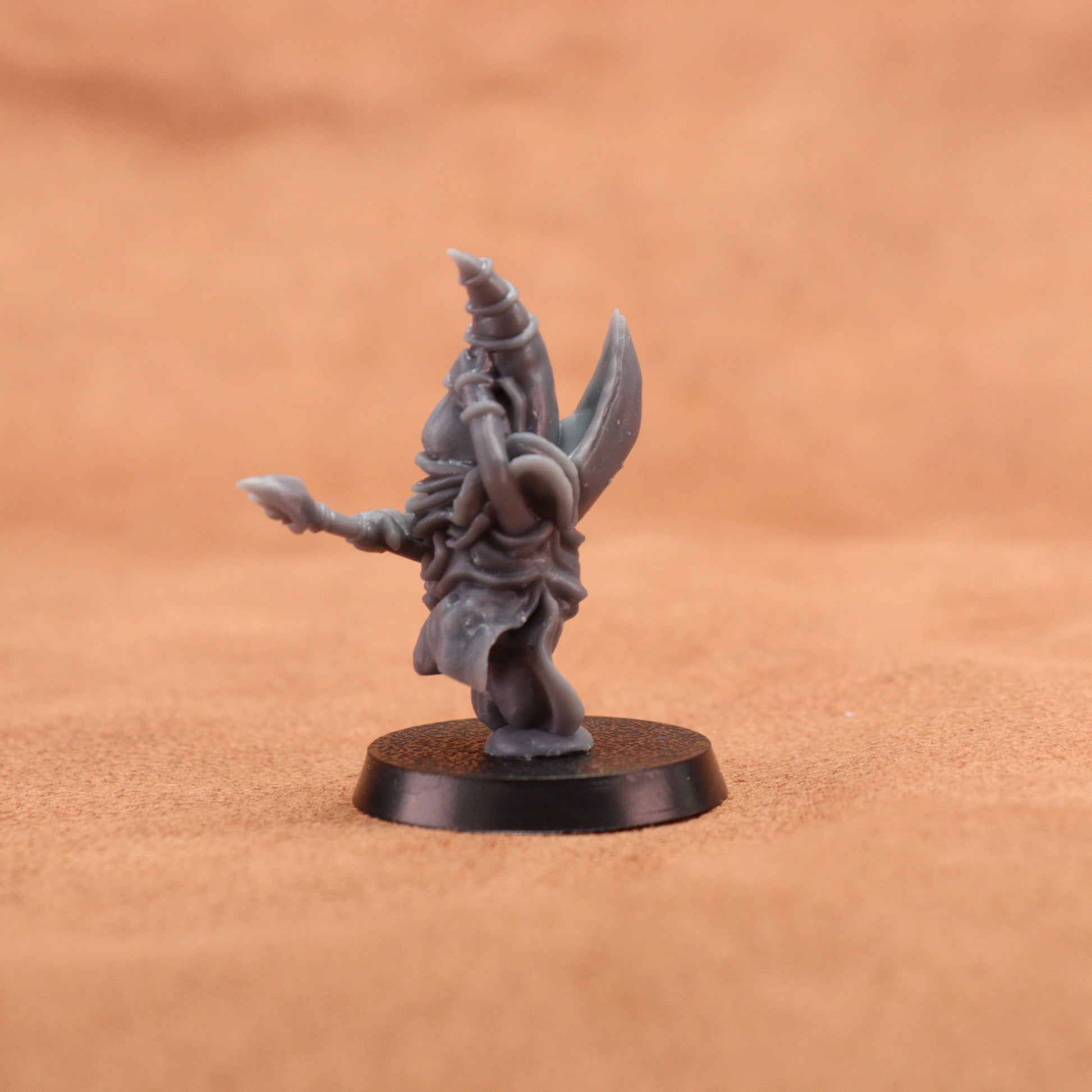 A small unpainted fantasy miniature figurine of a robed wizard or cleric holding a staff and shield, mounted on a round black base, photographed against a warm tan textured background.