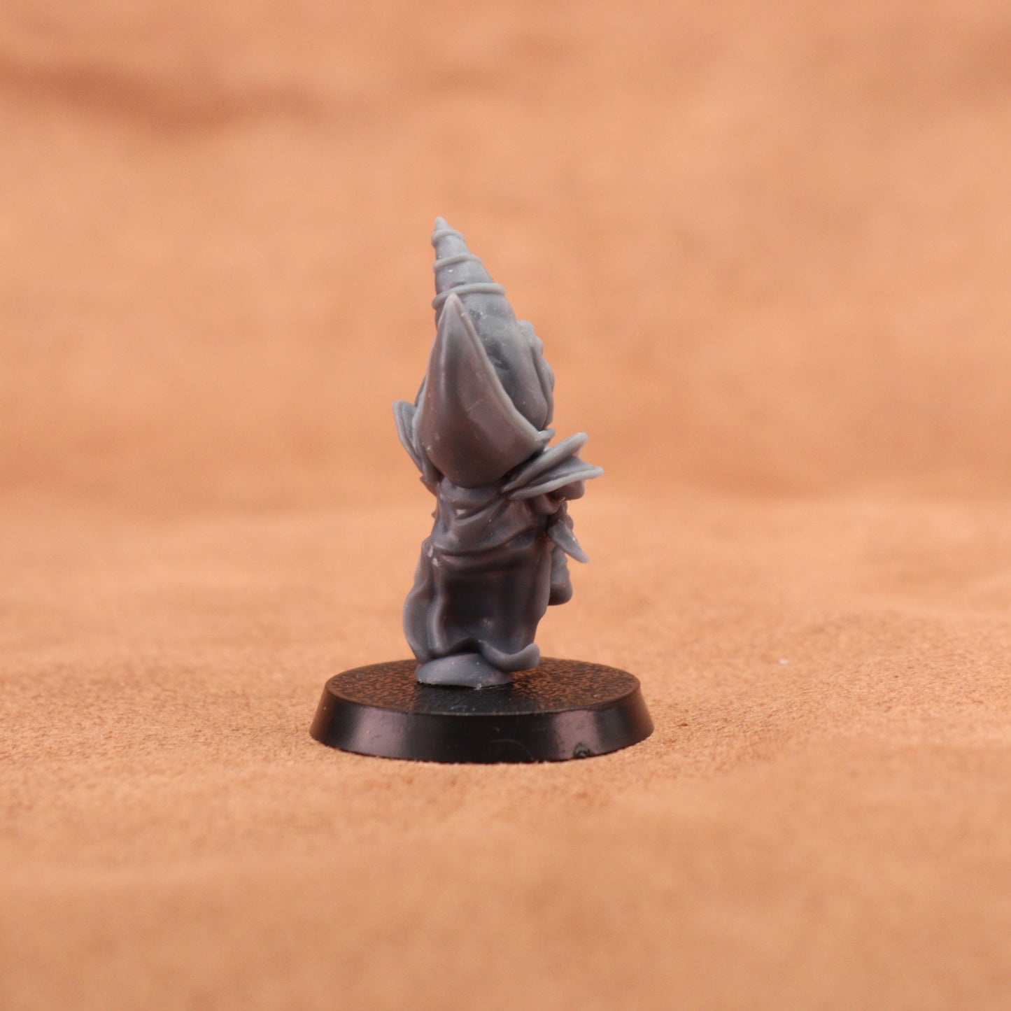 A small unpainted gray fantasy miniature figurine of a robed wizard or cleric standing on a round black base, photographed against a warm tan textured background.