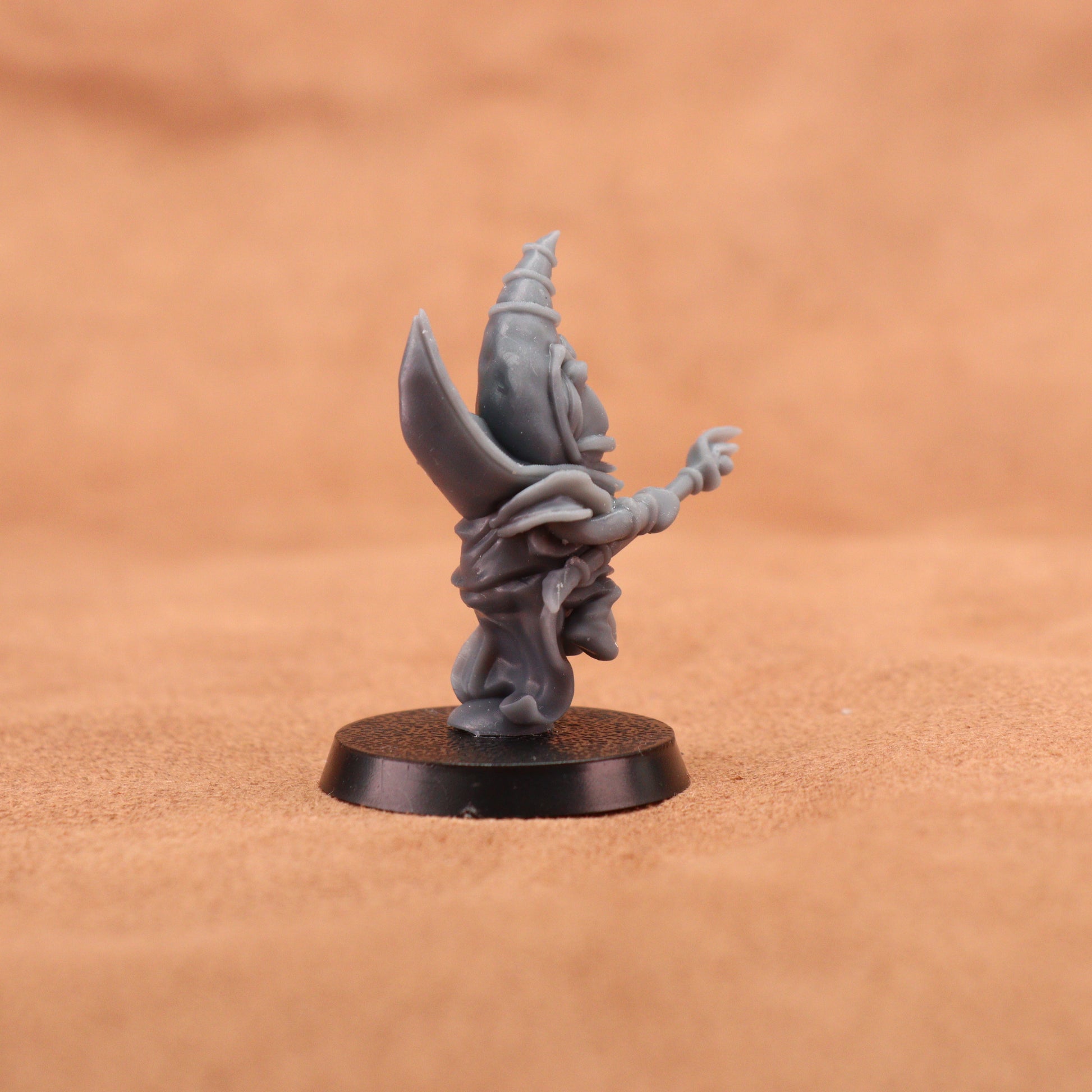 A small unpainted gray fantasy miniature figurine standing on a round black base, photographed against a warm tan textured background.