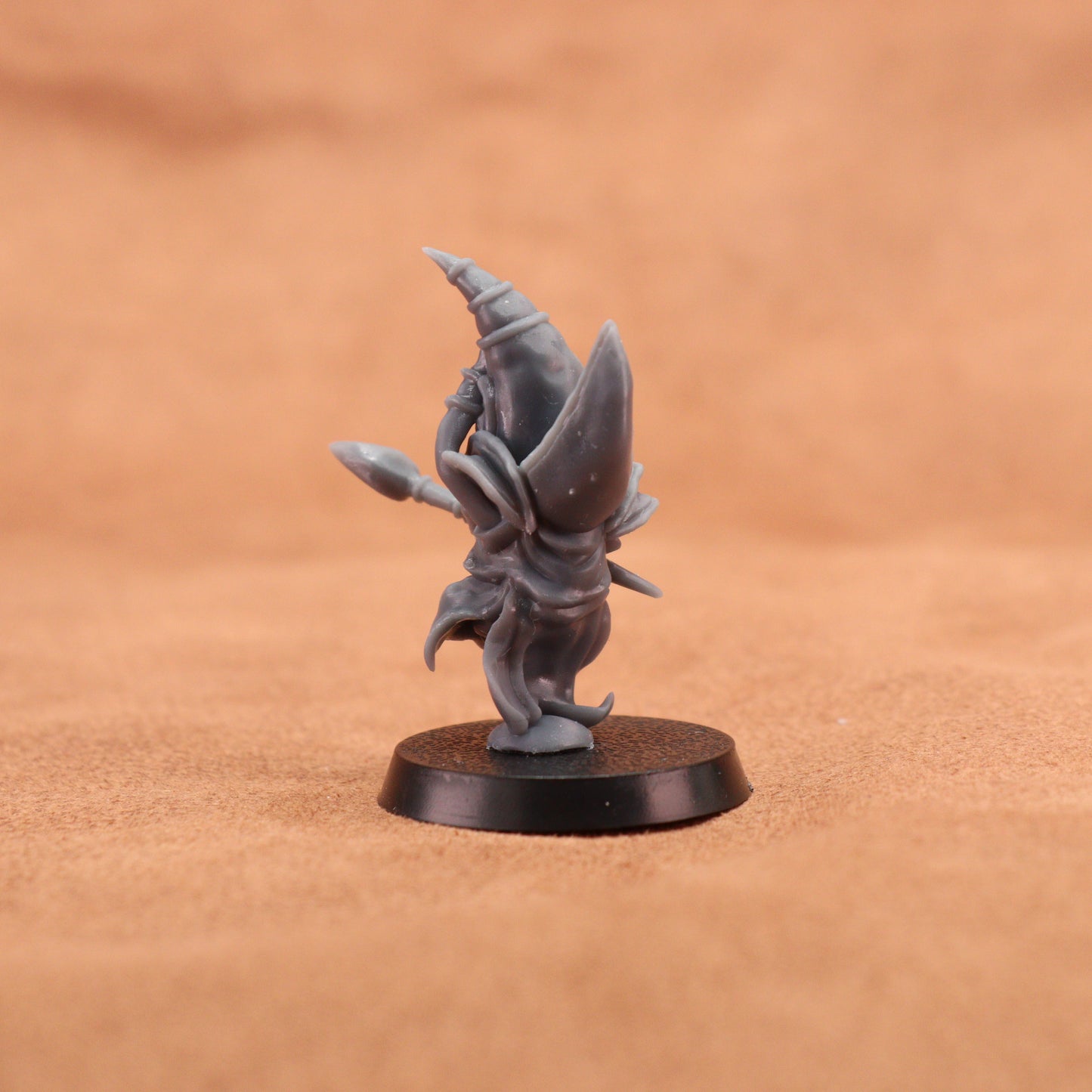 A small unpainted gray fantasy miniature figurine standing on a round black base, photographed against a warm tan textured background.
