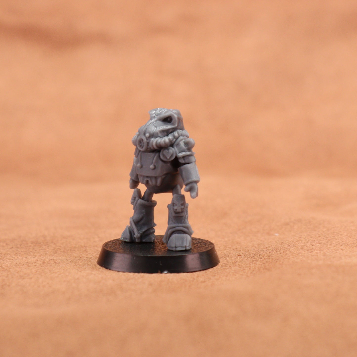 A small unpainted gray tabletop miniature figurine standing on a round black base, photographed against a warm tan textured background.