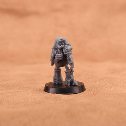 A small unpainted gray fantasy miniature figurine of a robotic or sci-fi armored character standing on a round black base, photographed against a warm tan textured background.