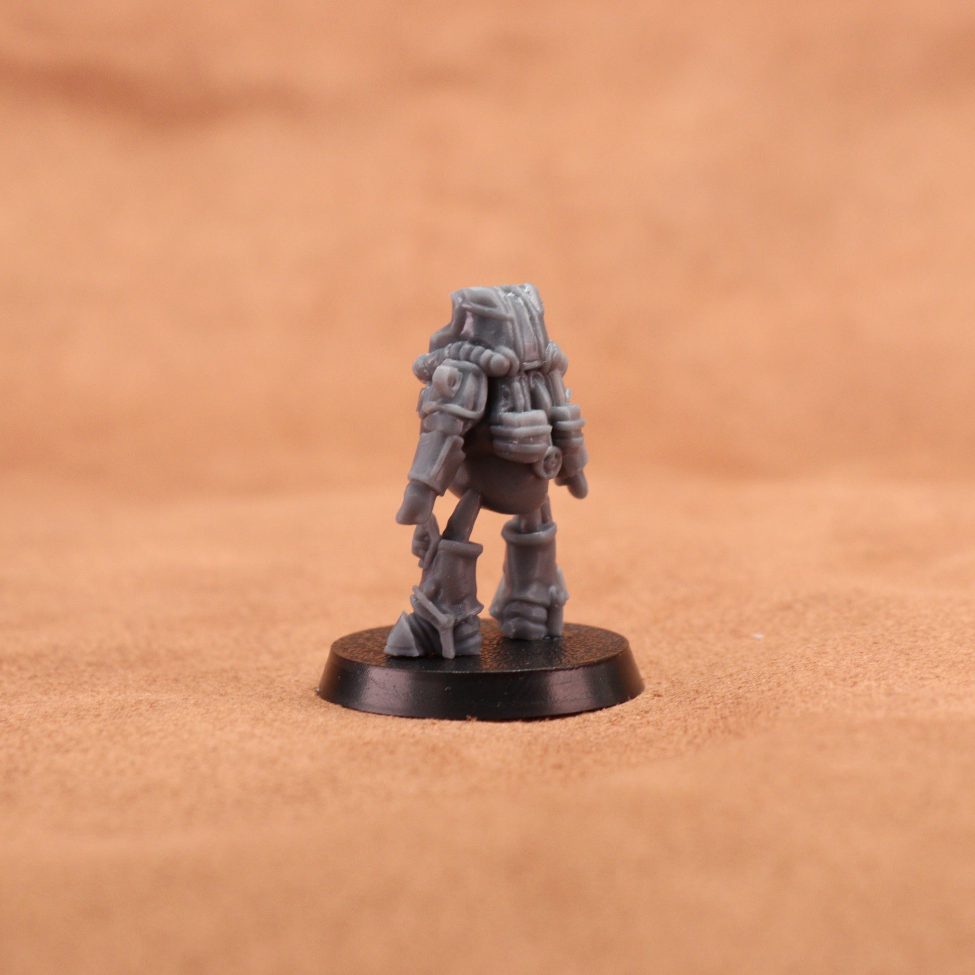 A small gray sci-fi miniature figurine standing on a round black base, photographed against a warm tan textured background.