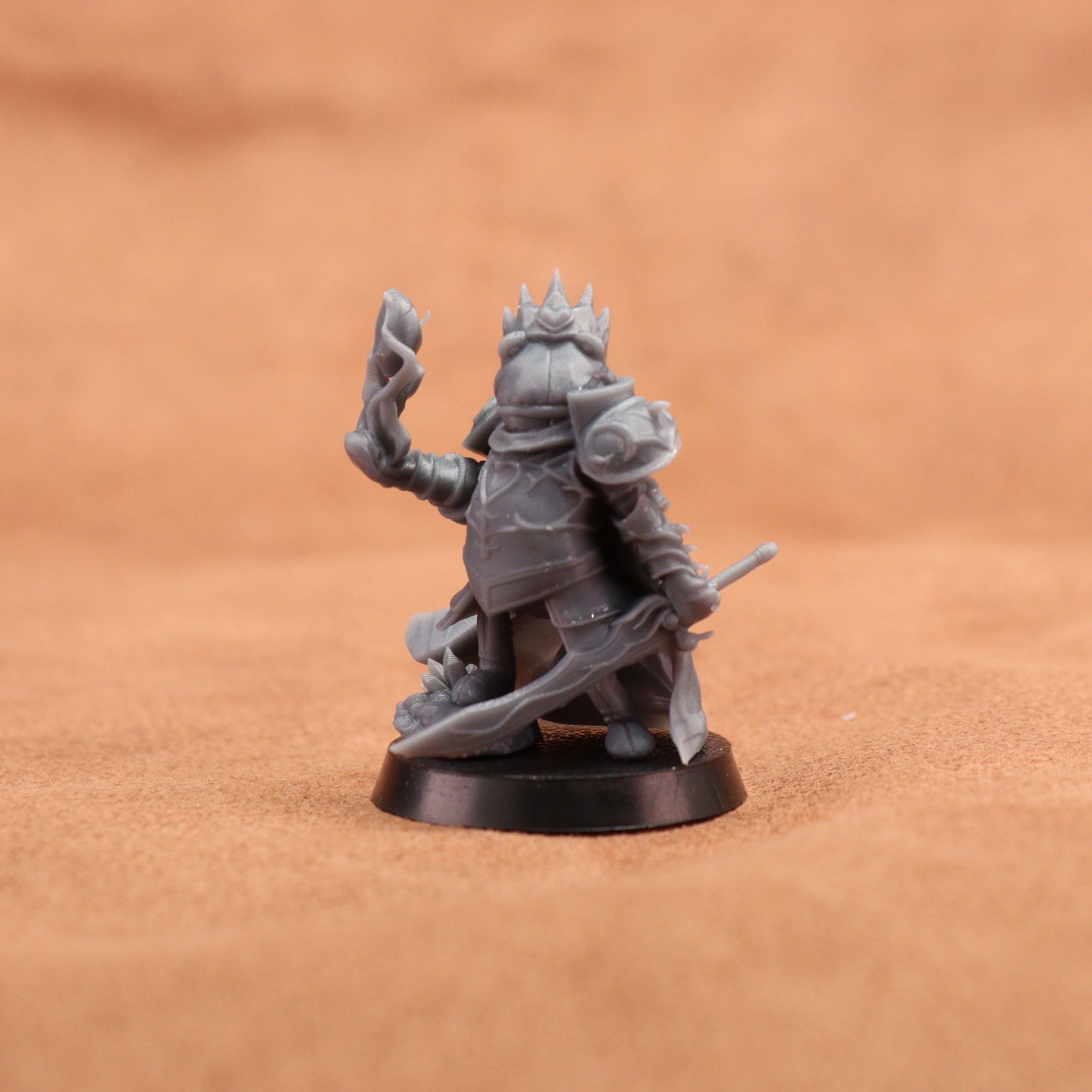 An unpainted gray fantasy miniature figurine of a crowned warrior holding a sword and staff, standing on a round black base against a warm brown textured background.