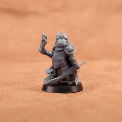 An unpainted gray fantasy miniature figurine of a crowned warrior holding a sword and staff, standing on a round black base against a warm brown textured background.
