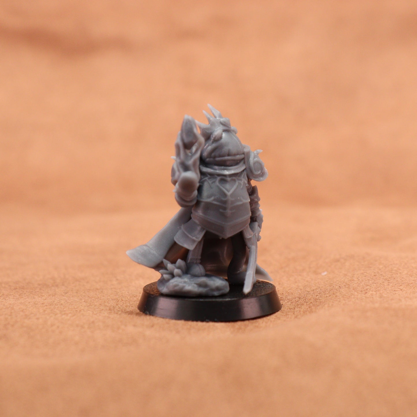 A small unpainted gray fantasy warrior miniature figurine standing on a round black base, photographed against a warm brown textured background.