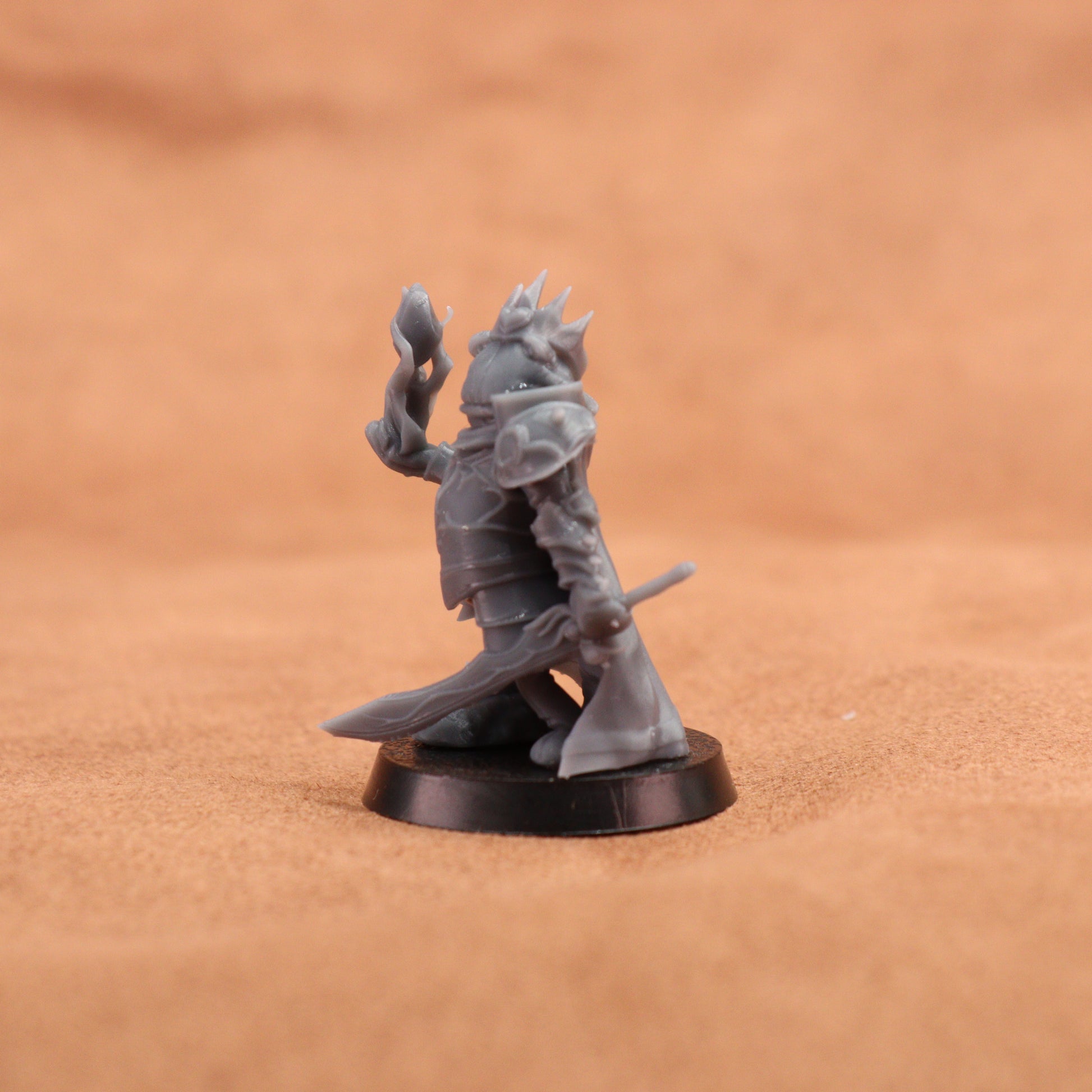 An unpainted gray fantasy miniature figurine of a crowned warrior holding a sword and staff, standing on a round black base against a warm brown textured background.