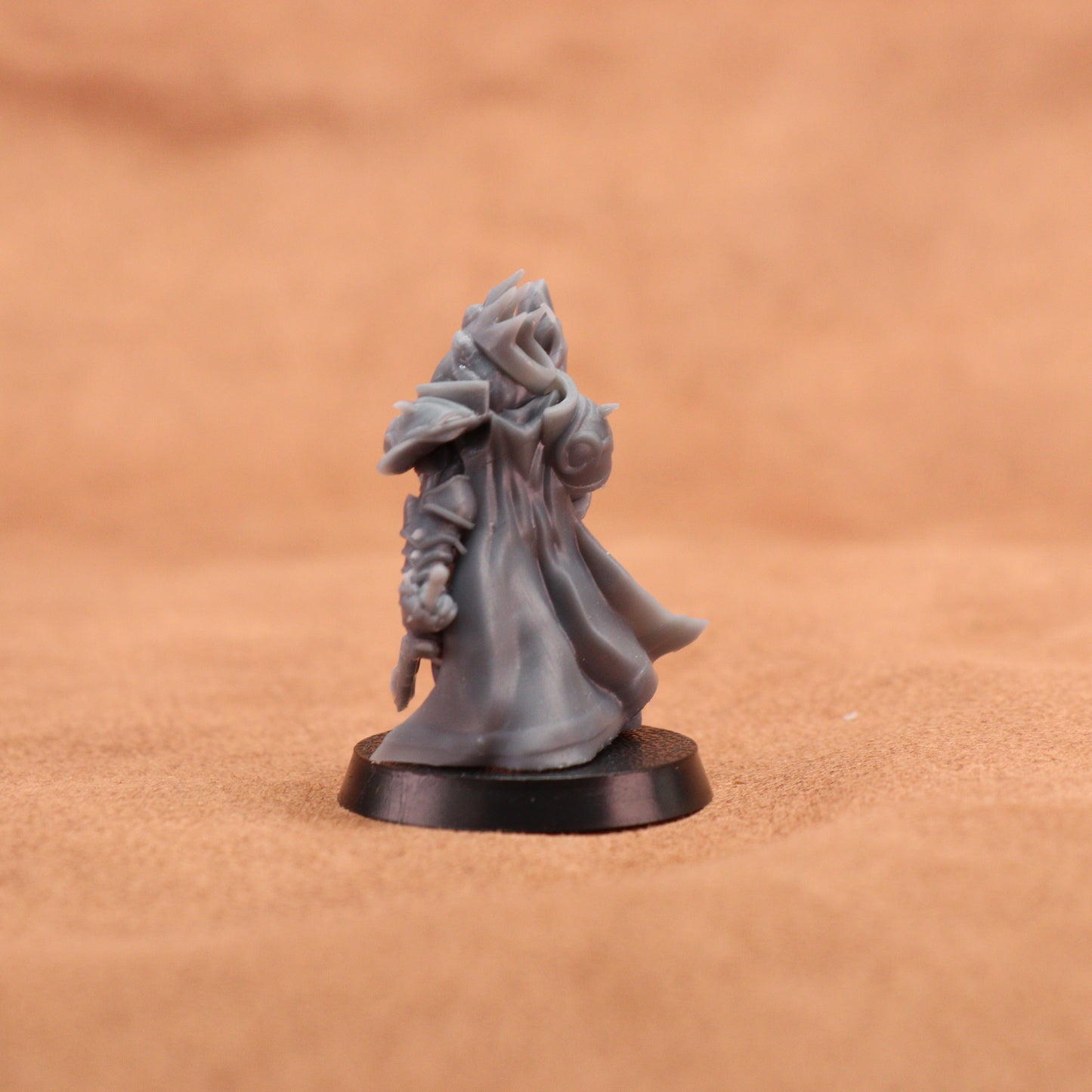 A small unpainted gray fantasy miniature figurine of a hooded robed character standing on a round black base, photographed against a warm tan textured background.