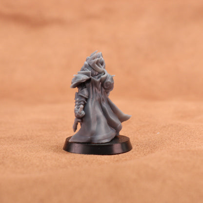 A small unpainted gray fantasy miniature figurine of a hooded robed character standing on a round black base, photographed against a warm tan textured background.