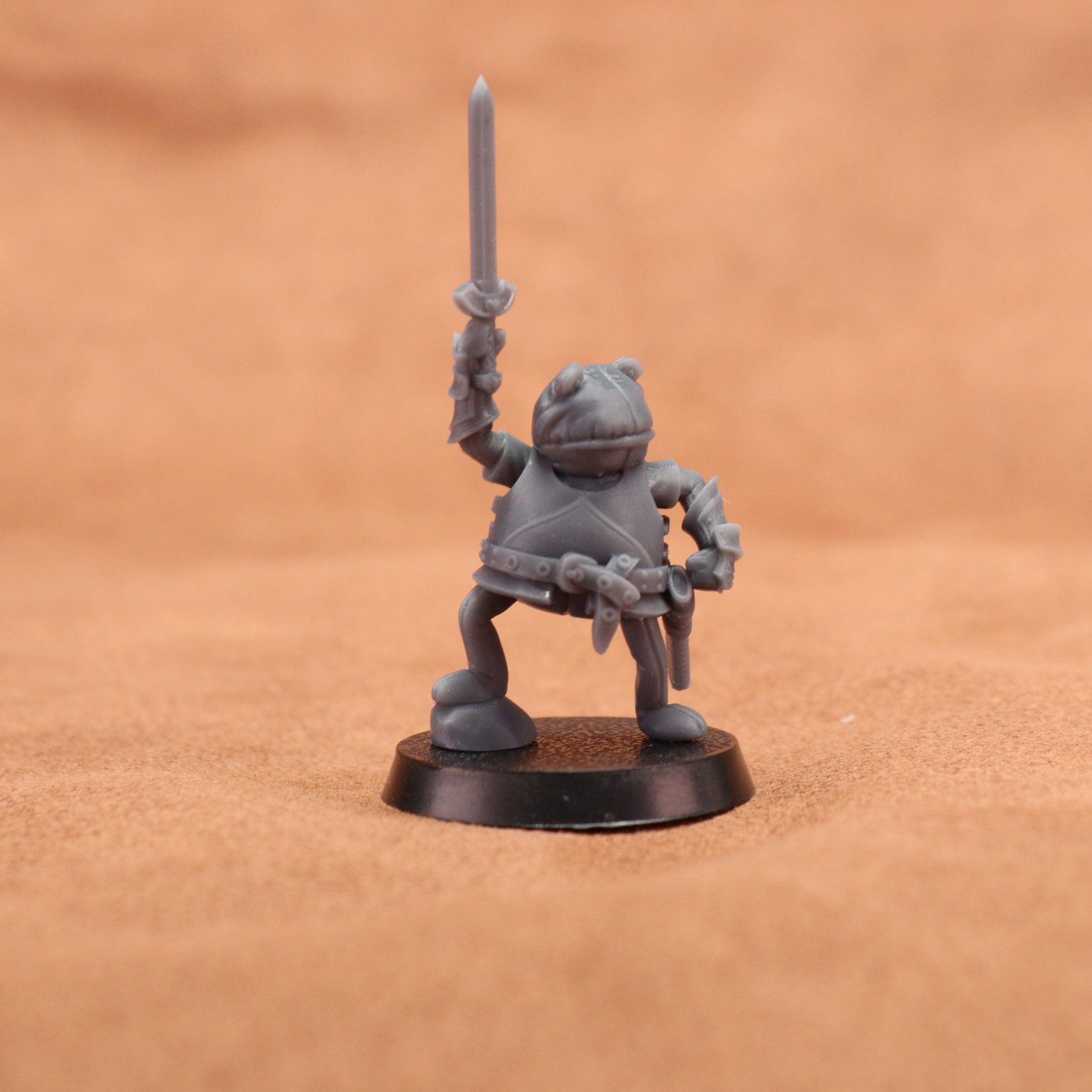 A small unpainted gray fantasy miniature figurine of a warrior holding a sword aloft, standing on a round black base against a warm brown textured background.