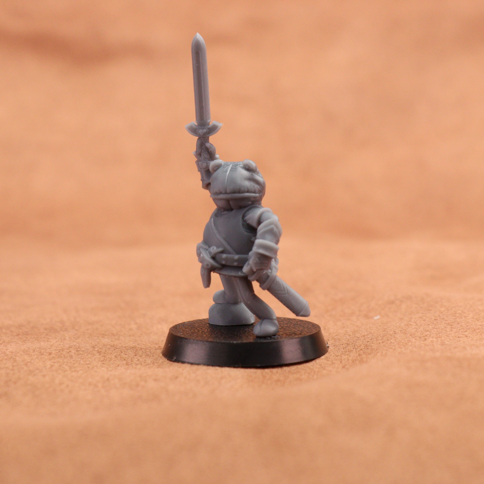 A small unpainted gray fantasy warrior miniature figurine holding a sword aloft, standing on a round black base against a warm sandy beige background.