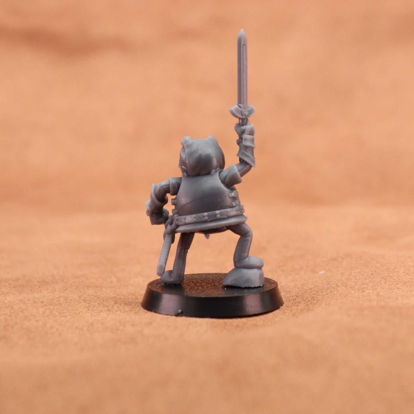 A small unpainted gray fantasy miniature figurine holding a sword aloft, standing on a round black base against a warm brown textured background.