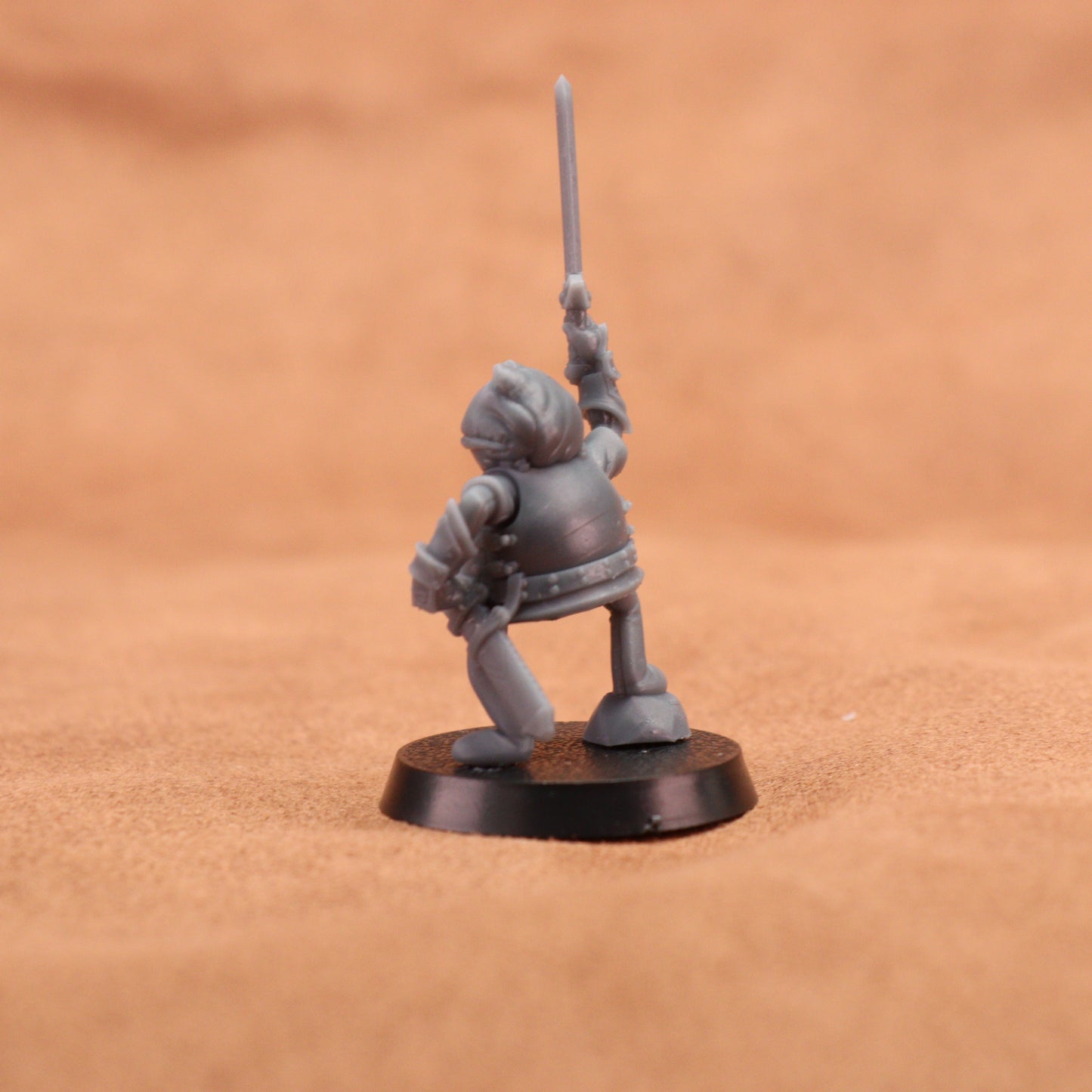 A small unpainted gray fantasy warrior miniature figurine holding a sword aloft, standing on a round black base against a warm brown textured background.