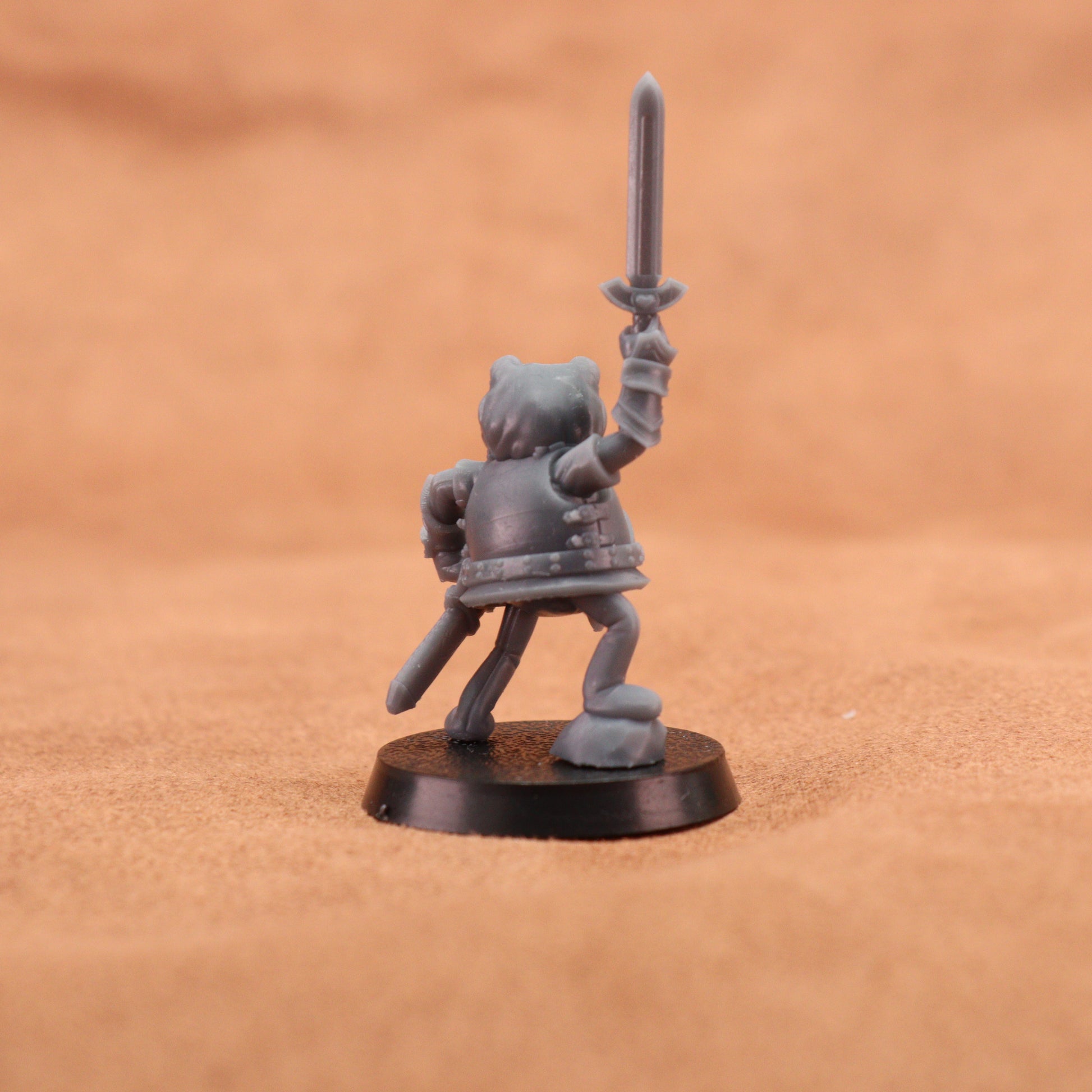 A small unpainted gray fantasy warrior miniature figurine holding a sword aloft, standing on a round black base against a warm tan textured background.