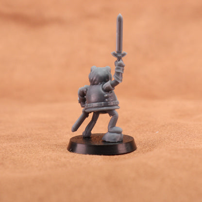A small unpainted gray fantasy warrior miniature figurine holding a sword aloft, standing on a round black base against a warm tan textured background.