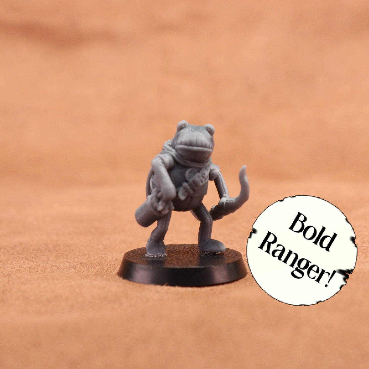 A small unpainted gray fantasy miniature figurine of a frog-like ranger standing on a round black base, photographed against a warm tan textured background. A white circular sticker with the text 'Bold Ranger!' is placed to the right.