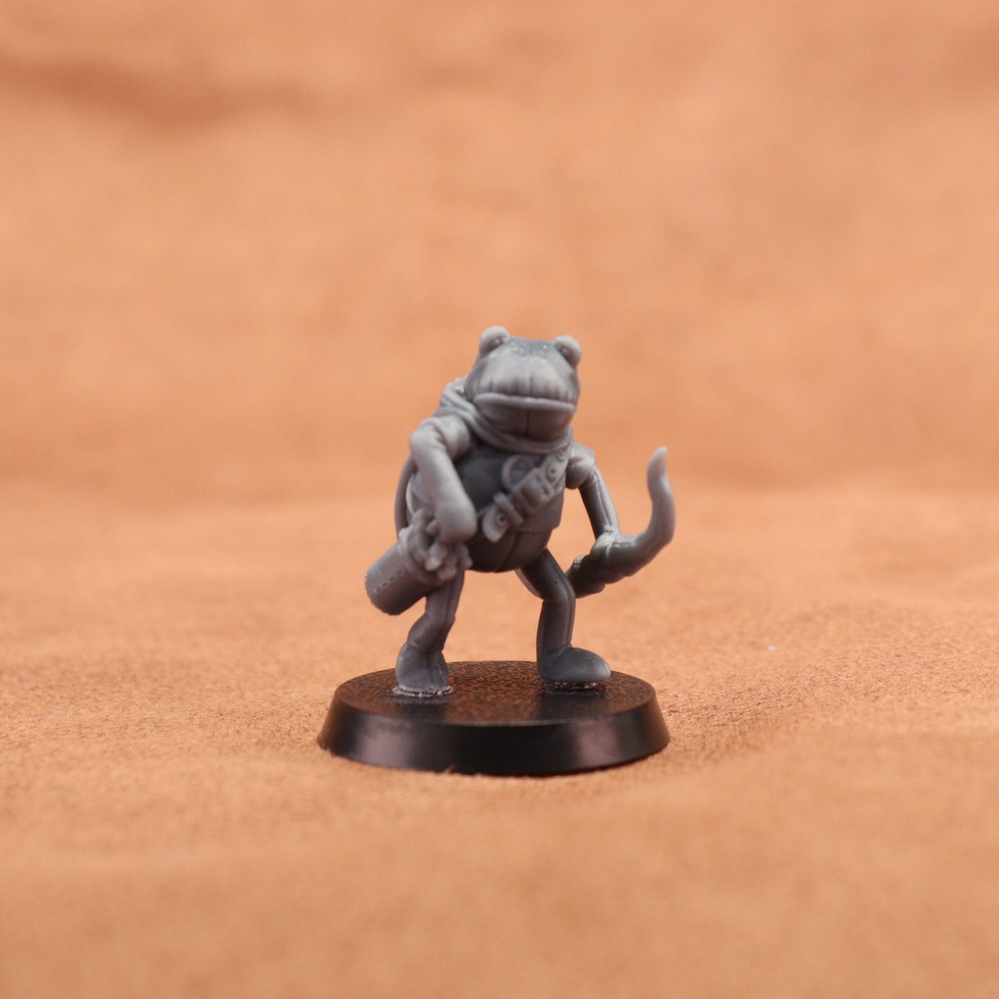 A small unpainted gray fantasy frog miniature standing on a round black base, photographed against a warm tan textured background.