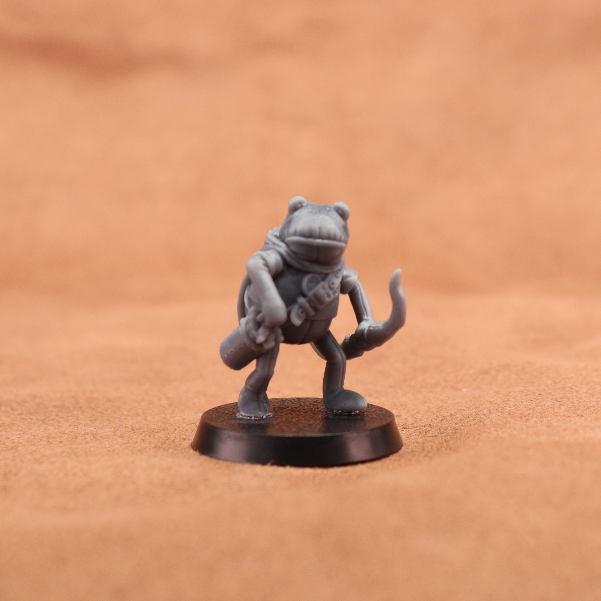 A small unpainted gray fantasy frog miniature standing on a round black base, photographed against a warm tan textured background.