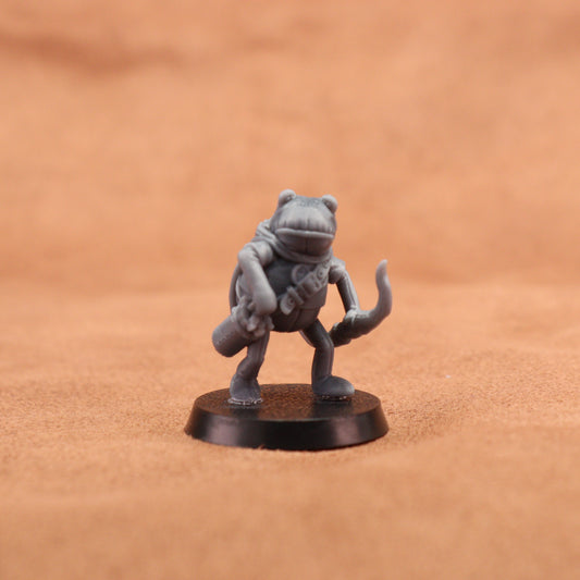 A small unpainted gray fantasy frog miniature standing on a round black base, photographed against a warm tan textured background.