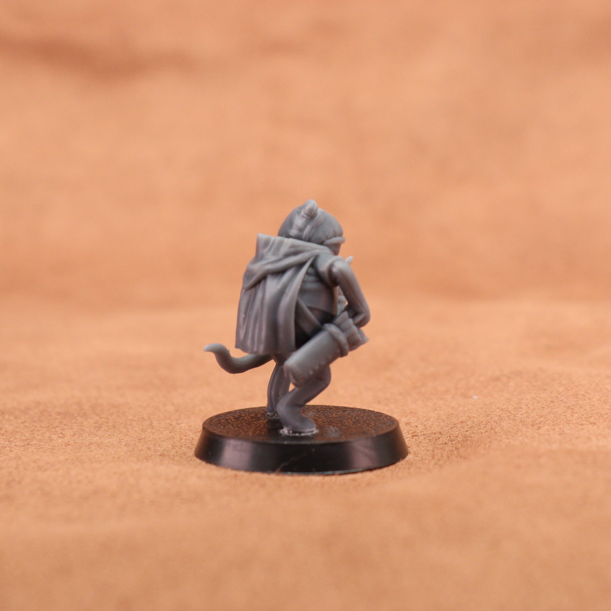 A small unpainted gray fantasy miniature figurine of a hooded creature crouching on a round black base, photographed against a warm brown textured background.