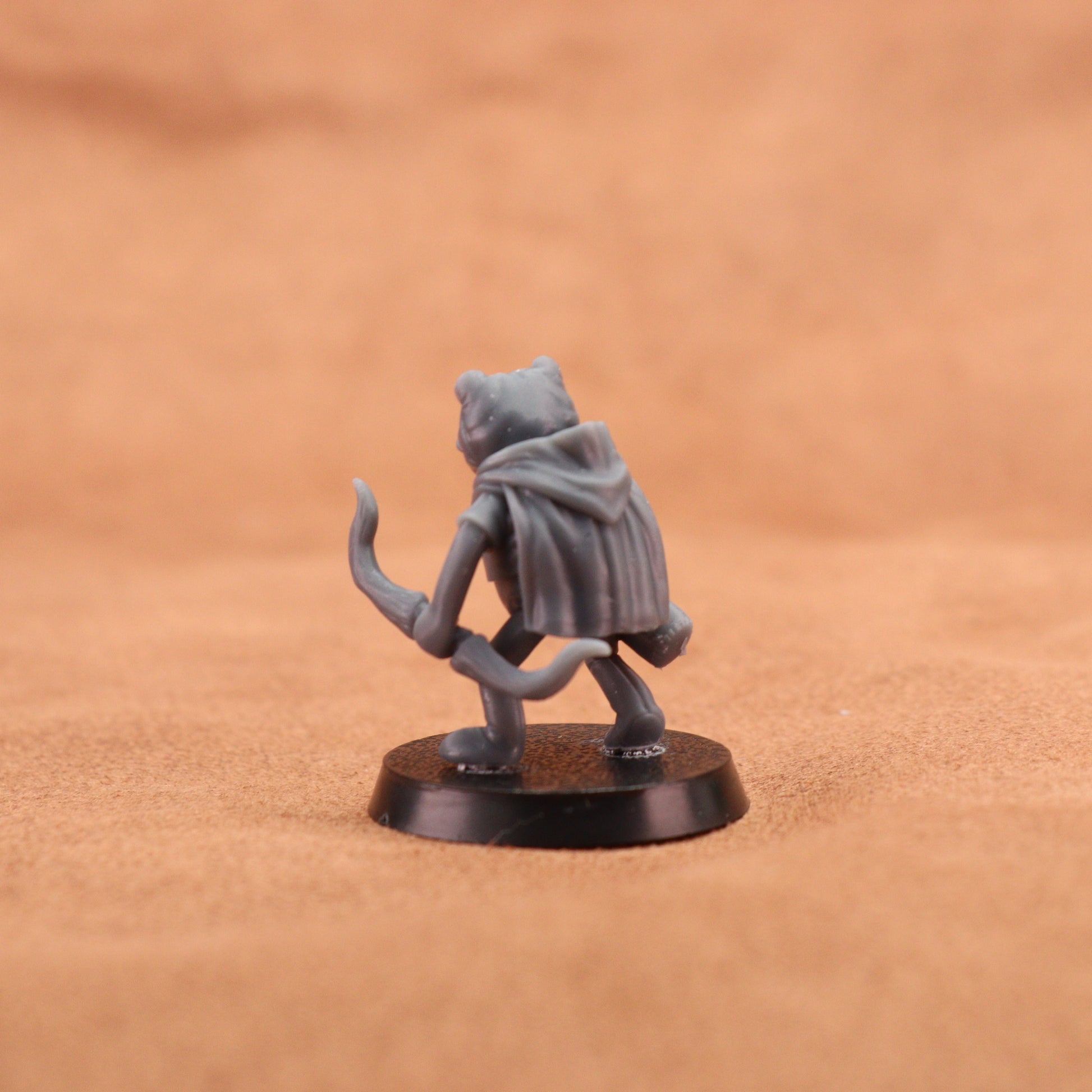 A small gray fantasy miniature figurine of a cat-like character wearing a hooded cloak, standing on a round black base against a warm tan textured background.