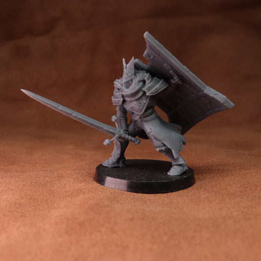 A gray unpainted fantasy warrior miniature figurine holding a sword and shield, standing on a round black base against a brown textured background.