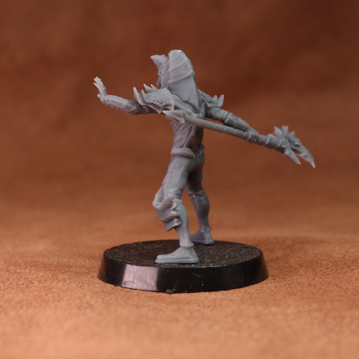 A gray unpainted fantasy miniature figurine of a warrior holding a spear, standing on a round black base against a textured brown background.