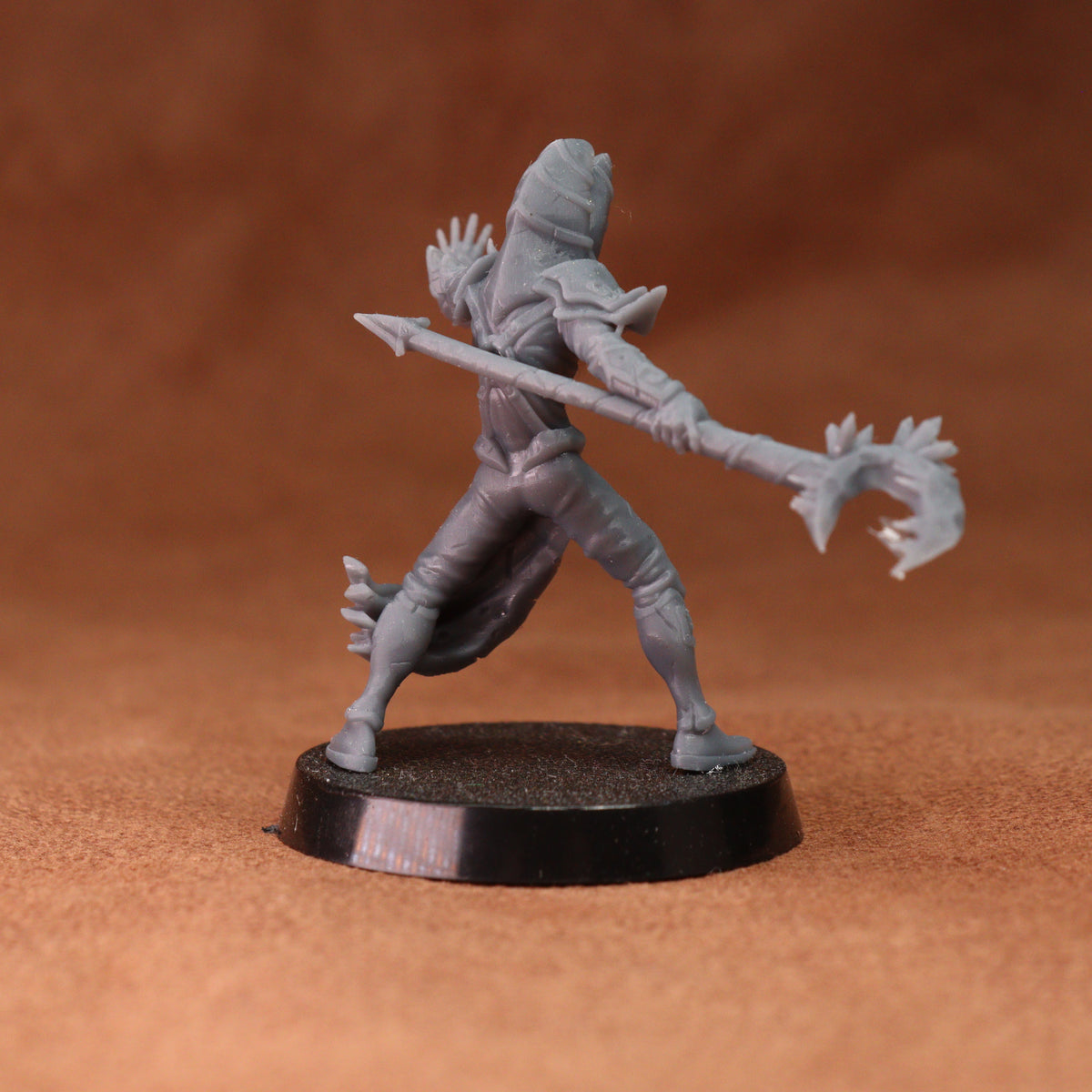 An unpainted gray fantasy miniature figurine of a hooded warrior holding a spear, standing on a round black base against a textured brown background.