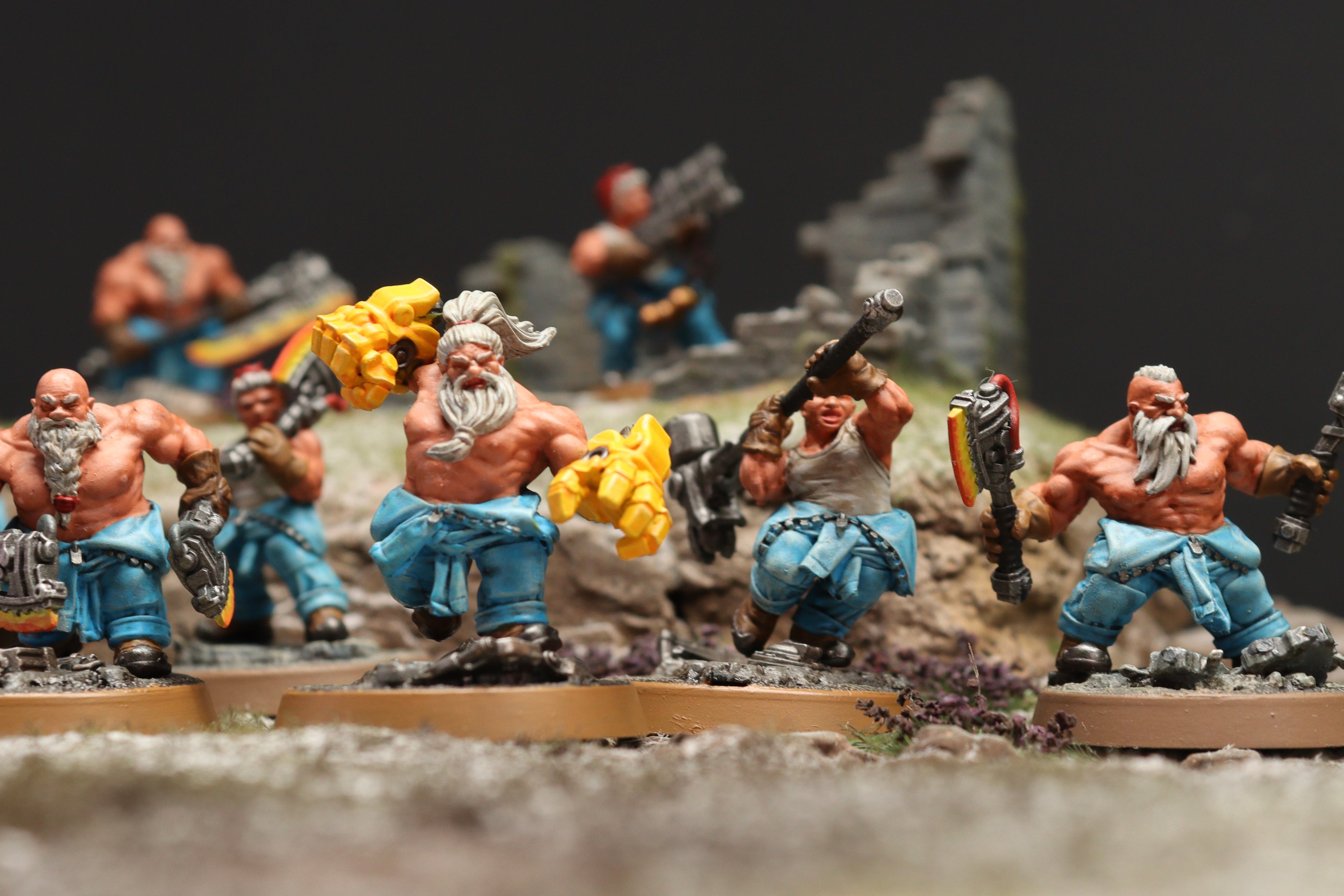 A group of painted fantasy warrior miniatures posed on a tabletop diorama with rocky terrain and a dark backdrop. The figures are muscular, mostly shirtless, with blue pants, gray hair/beards, and various weapons including maces and shields.