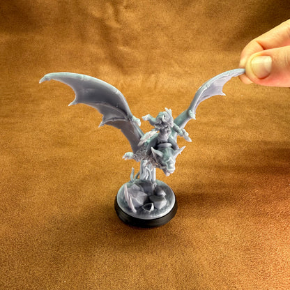 Miniature dragon figurine on a brown surface with a hand holding its wing.