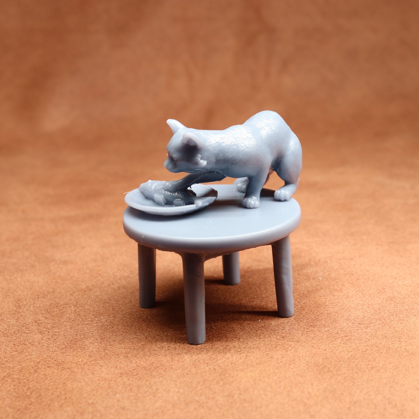 Miniature figurine of a cat on a small table with a fish, all in a light gray color, on a brown background.