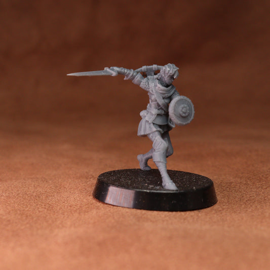 An unpainted gray plastic miniature figurine of a warrior with a sword and shield, standing on a round black base against a textured brown background.