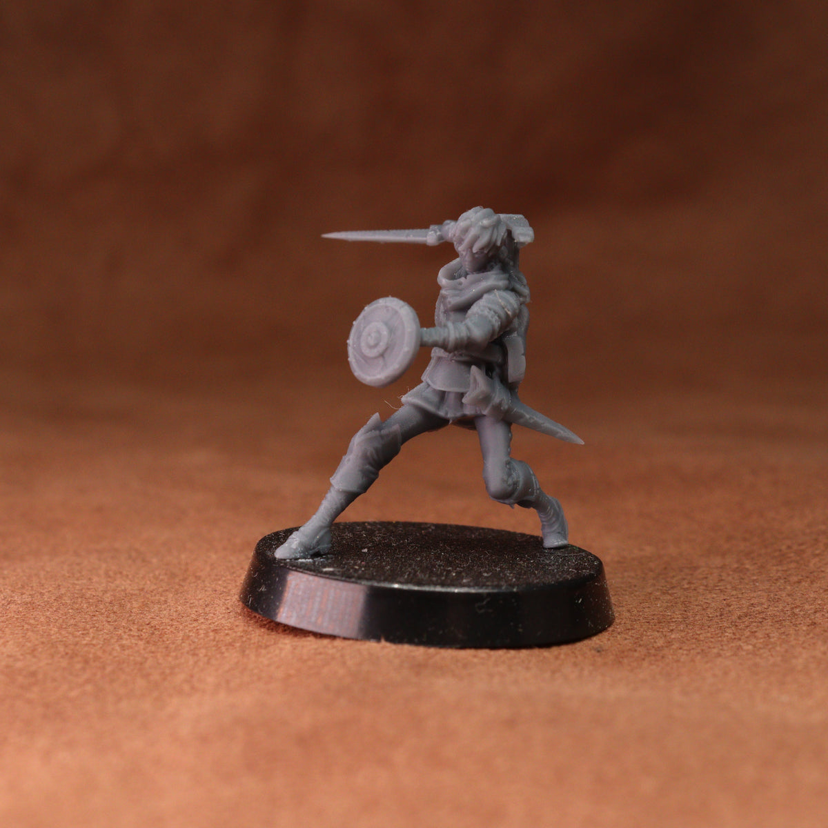 An unpainted gray fantasy warrior miniature figurine holding a sword and shield, standing on a round black base against a textured brown background.