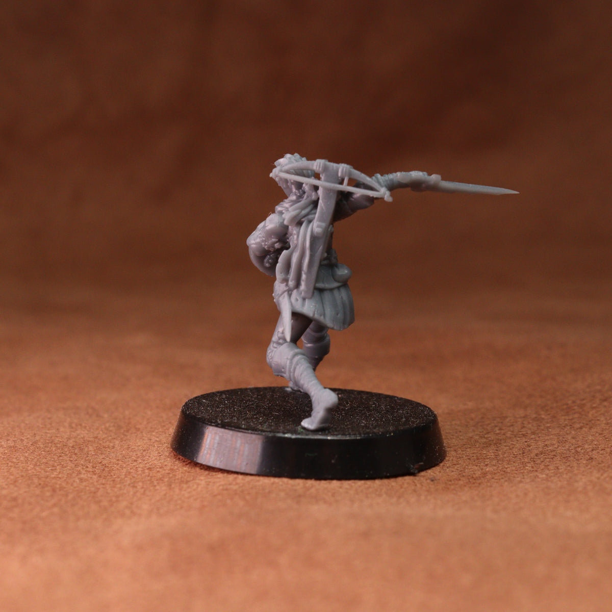 An unpainted gray plastic miniature figurine of a warrior holding a spear, standing on a round black base against a textured brown background.