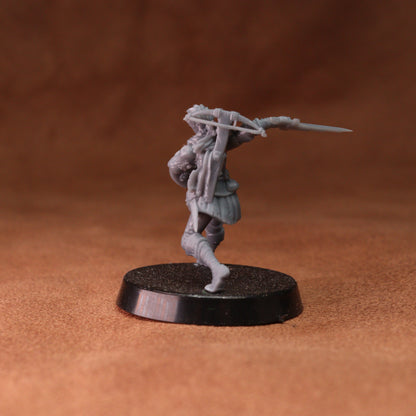 An unpainted gray plastic miniature figurine of a warrior holding a spear, standing on a round black base against a textured brown background.